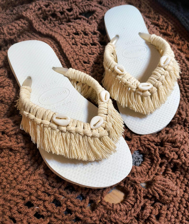 May include: A pair of off-white flip-flops with macrame detailing. The straps are adorned with beige fringe and small seashells. The flip-flops are displayed on a brown crocheted surface.