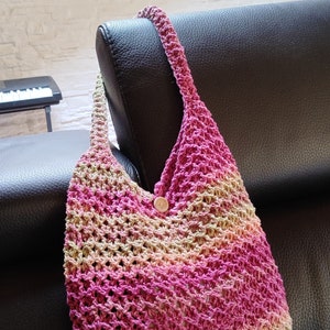 May include: A pink and beige crocheted tote bag with a wooden button. The bag is hanging on the back of a black leather couch.