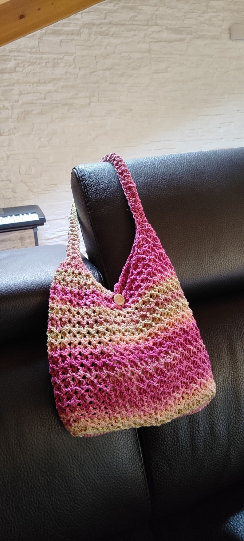 May include: A pink and beige crocheted tote bag with a wooden button. The bag is hanging on the back of a black leather couch.