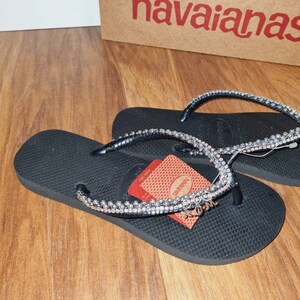 May include: Black flip-flops with a rhinestone strap and a "Mom" charm. The flip-flops are on a wooden surface. The flip-flops are in a box that says "havaianas".