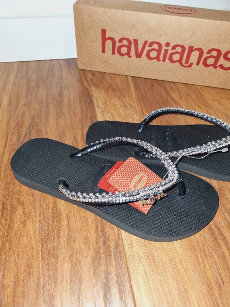 May include: Black flip-flops with a rhinestone strap and a "Mom" charm. The flip-flops are on a wooden surface. The flip-flops are in a box that says "havaianas".