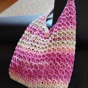 May include: A pink and beige crocheted tote bag with a wooden button. The bag is hanging on a black leather chair.