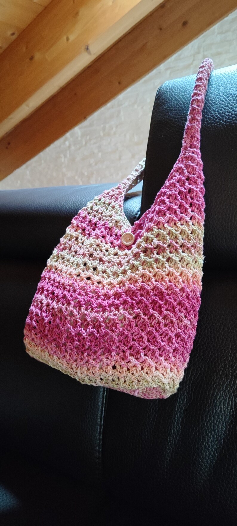 May include: A pink and beige crocheted tote bag with a wooden button. The bag is hanging on a black leather chair.