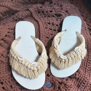 May include: A pair of white Havaianas flip-flops with beige macrame detailing. The macrame features fringe and small seashells. The flip-flops are displayed on a brown, textured surface.