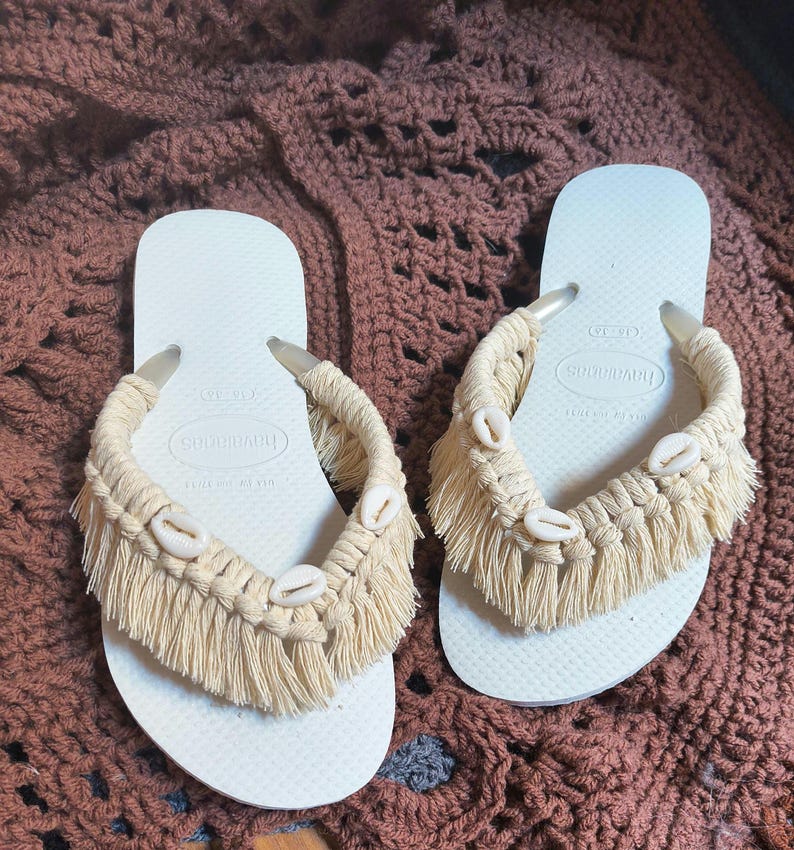 May include: A pair of white Havaianas flip-flops with beige macrame detailing. The macrame features fringe and small seashells. The flip-flops are displayed on a brown, textured surface.