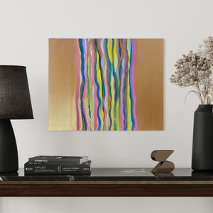 Puede incluir: Pintura abstracta con líneas onduladas de colores sobre un fondo dorado. La pintura está colgada en una pared blanca sobre una mesa de mármol negro con una lámpara y un jarrón.