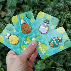 May include: Four enamel pins featuring cartoon animal characters on a green and yellow background. The pins are on cards with the text "Newloot" printed on them. The characters are a beaver wearing a party hat, a penguin wearing a top hat, a turtle wearing a hat, and a duck wearing a bow.