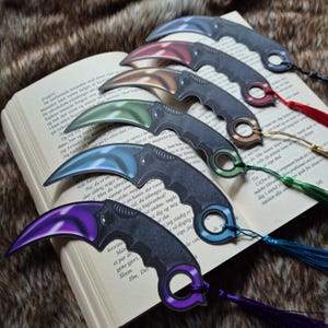 Könnte beinhalten: Eine Sammlung von sechs Lesezeichen im Karambit-Stil mit gebogenen Klingen in verschiedenen Farben, darunter Lila, Blau, Grün, Gold, Rot und Grau. Jedes Lesezeichen hat eine passende Quaste. Die Lesezeichen liegen auf einem offenen Buch.