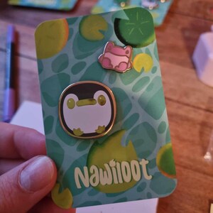 May include: A green and white card with a penguin and a frog pin on it. The card has the text "Nawilooot" on it.