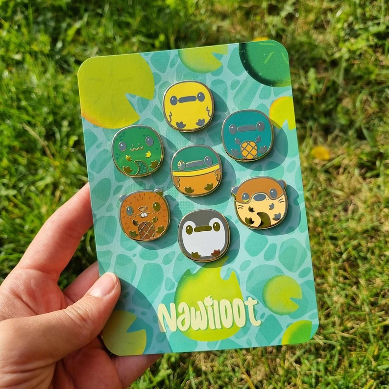 May include: A set of eight enamel pins featuring various aquatic creatures, including a turtle, frog, otter, and penguin. The pins are colorful and have a cartoonish style. The background is a green and blue pattern with the word "Nawilioot" printed in white.
