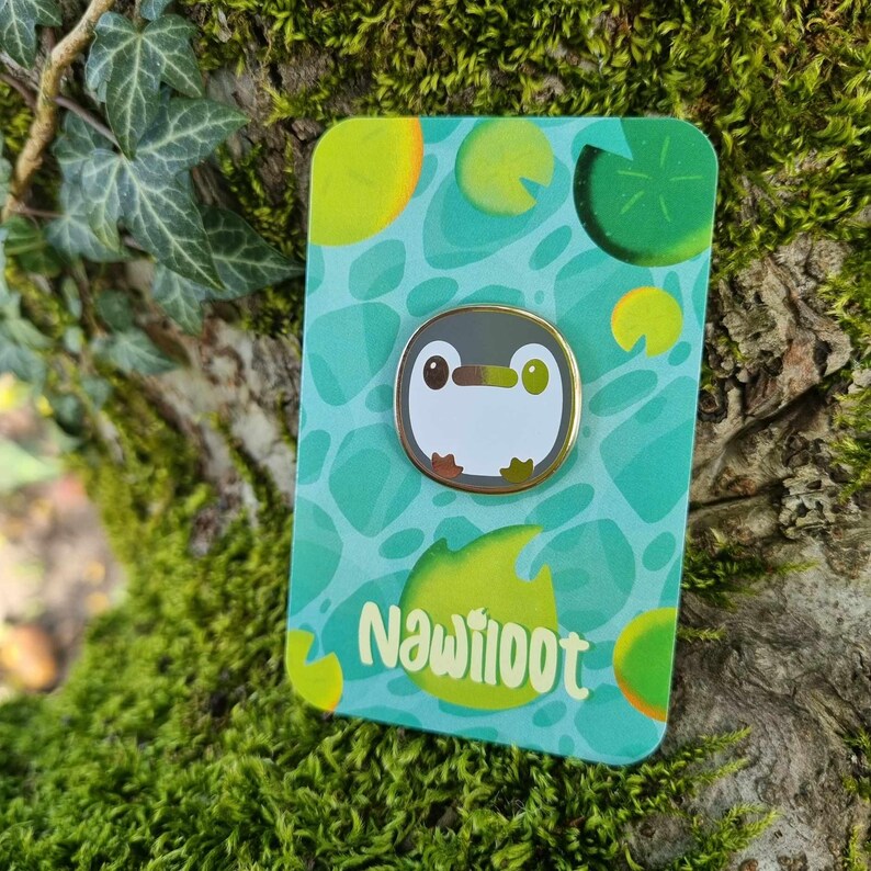 May include: A gold-colored enamel pin featuring a cartoon penguin with a white body, black eyes, and a yellow beak. The pin is set against a green and yellow background with a pattern of lily pads and the text "Nawiiioot".