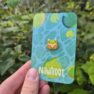 May include: A small enamel pin featuring a cartoon frog design in green, orange, and beige. The pin is attached to a teal card with a lily pad pattern and the word "Nawiloot" in white. The background is blurred green foliage.