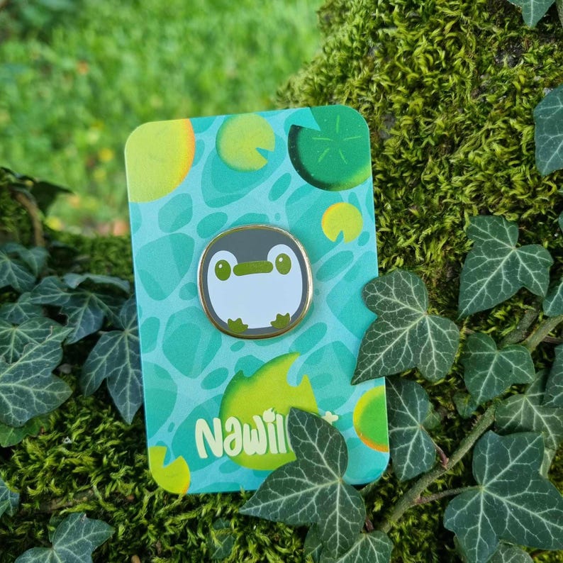 May include: A green, blue, and yellow enamel pin featuring a cartoon penguin with a white body, black eyes, and a green beak. The pin is on a card with the word "Nawil" printed on it.