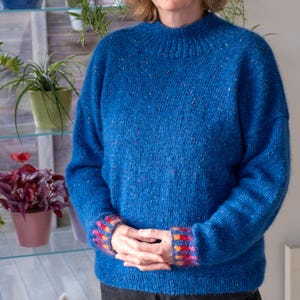 May include: A blue knit sweater with a high neckline and speckled detailing. The cuffs feature a colorful pattern of red, orange, and yellow squares. The sweater is a medium weight and appears to be handmade.