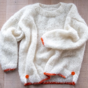 May include: A cream-colored, oversized sweater with a crew neck and long sleeves. The cuffs and hem are trimmed with bright orange yarn. Two orange buttons are visible near the hem. The sweater is made of a soft, fluffy yarn.