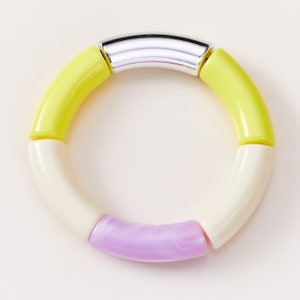 Könnte beinhalten: Ein farbenfrohes Armband mit Segmenten in Gelb, Weiß, Lavendel und einem silberfarbenen Metallverschluss. Das Armband besteht aus gebogenen, zylindrischen Perlen. Das Design ist modern und auffällig, geeignet für den Alltag oder besondere Anlässe.