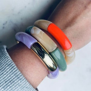 Könnte beinhalten: Ein Set aus vier bunten Armreifen. Die Armreifen sind auf dem Handgelenk einer Person gestapelt. Die Armreifen bestehen aus verschiedenen Materialien, darunter Kunststoff, Metall und Holz. Die Armreifen sind in verschiedenen Farben, darunter Lila, Orange, Grün und Creme.