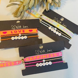 Könnte beinhalten: Drei Armband-Sets auf schwarzen Karten. Jedes Set enthält ein Armband mit einem positiven Wort wie "LOVE" oder "HAPPINESS" und ein Perlenarmband. Die Armbänder sind in Rosa, Orange, Gelb und Weiß gehalten.