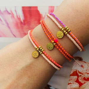 Könnte beinhalten: Vier farbige Perlenarmbänder mit goldenen Anhängern am Handgelenk einer Person. Die Armbänder sind rosa, orange und lila.