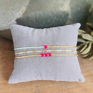 Könnte beinhalten: Drei Perlenarmbänder aus einer Kombination von metallisch goldfarbenen Saatperlen und hellen mintgrünen Saatperlen. Das oberste Armband hat eine leuchtend rosa Perle, das mittlere zwei und das untere Armband drei leuchtend rosa Perlen.