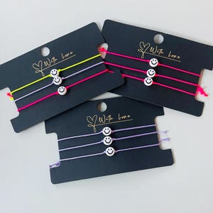 Könnte beinhalten: Drei Armband-Sets auf schwarzen Karten. Jedes Armband hat eine Smiley-Gesicht-Perle und farbiges Garn in Gelb, Pink und Lila. Auf den Karten steht in goldener Schrift "With love".