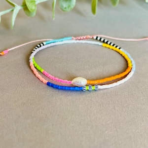 Könnte beinhalten: Ein farbenfrohes Perlenarmband mit einem weißen Perlenanhänger. Das Armband besteht aus verschiedenen Farben, darunter Blau, Grün, Pink, Orange und Weiß.