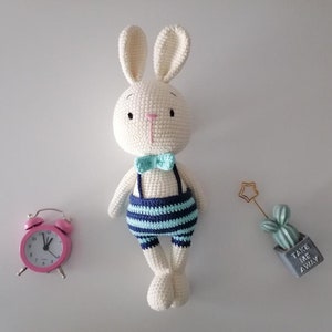 May include: A white crocheted bunny wearing a blue and white striped outfit and a light blue bow tie. The bunny is standing on a white surface with a pink alarm clock and a small potted cactus with the text "Take me away" on the pot.
