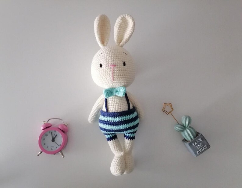 May include: A white crocheted bunny wearing a blue and white striped outfit and a light blue bow tie. The bunny is standing on a white surface with a pink alarm clock and a small potted cactus with the text "Take me away" on the pot.