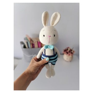 May include: A white crocheted bunny wearing blue and light blue striped overalls with a light blue bow tie.