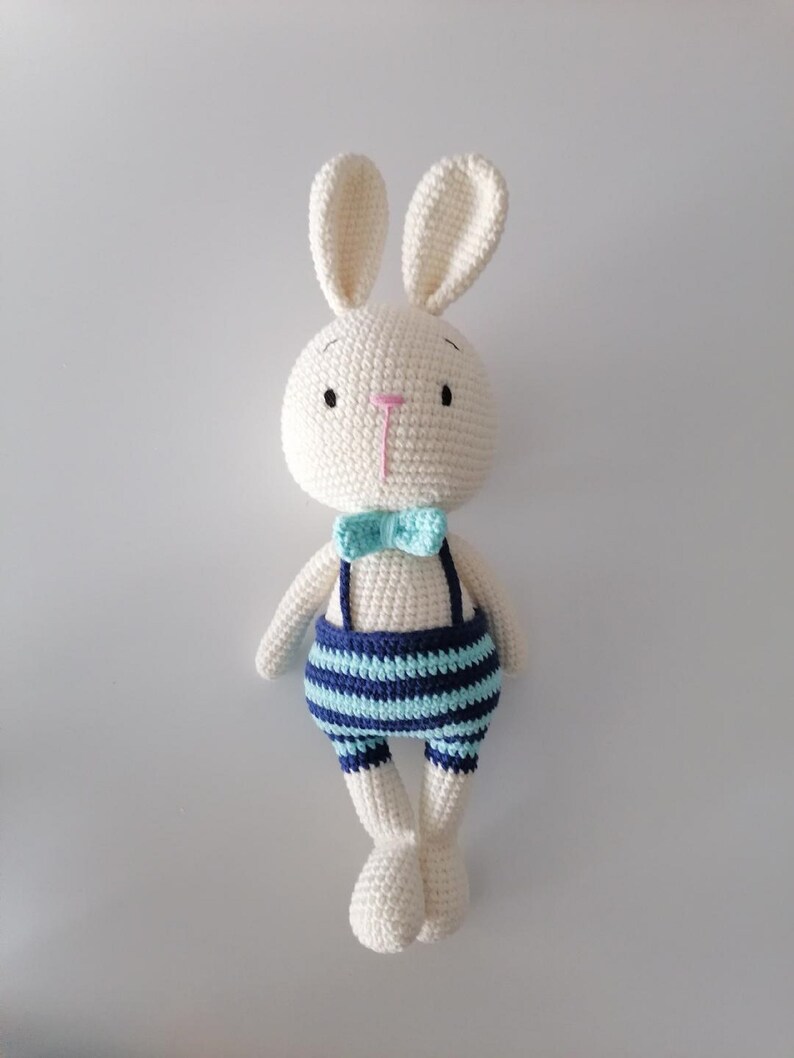 May include: White crocheted bunny rabbit wearing blue and turquoise striped overalls with a turquoise bow tie.