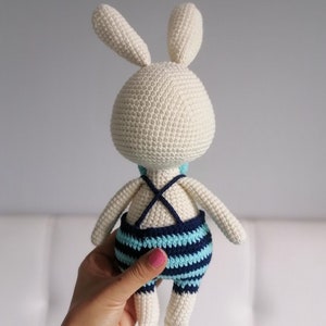 May include: A white crocheted bunny wearing a blue and turquoise striped swimsuit. The bunny is being held by a hand.