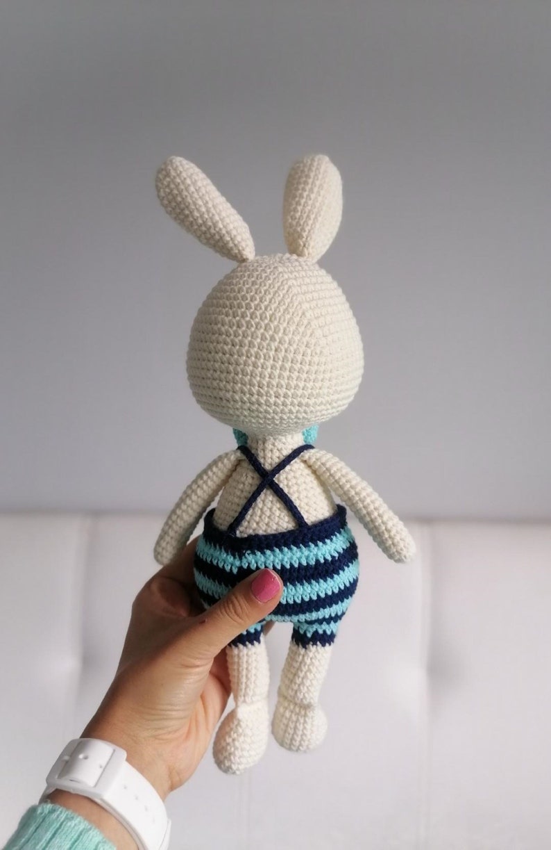 May include: A white crocheted bunny wearing a blue and turquoise striped swimsuit. The bunny is being held by a hand.