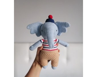 May include: A blue crocheted elephant toy wearing a red and blue striped shirt, brown pants, and a blue and red hat.