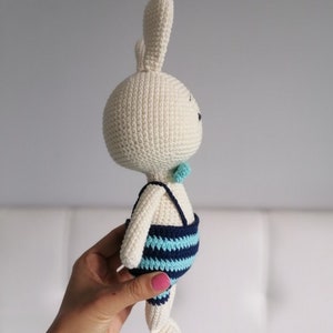 May include: A white crocheted bunny wearing a blue and turquoise striped swimsuit. The bunny is holding a small turquoise bow tie.
