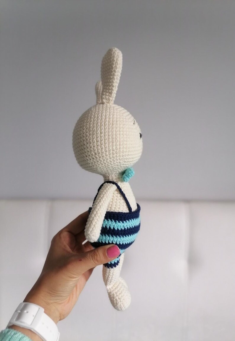 May include: A white crocheted bunny wearing a blue and turquoise striped swimsuit. The bunny is holding a small turquoise bow tie.