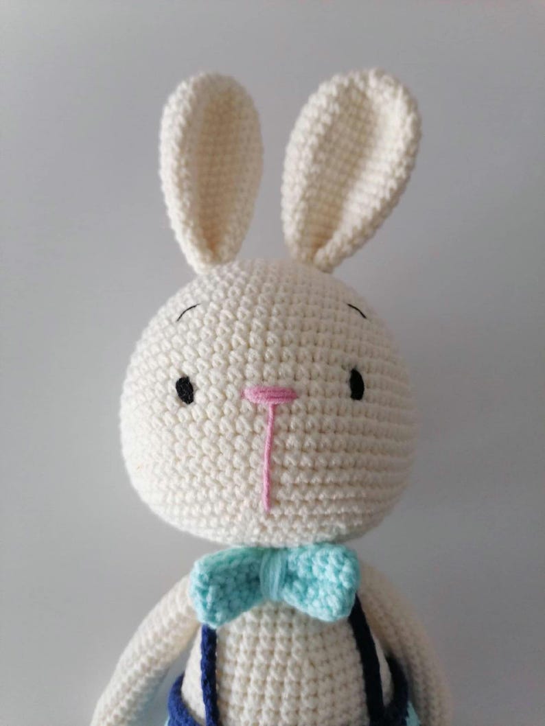 May include: A white crocheted bunny with a blue and turquoise bow tie. The bunny has black eyes and a pink nose.