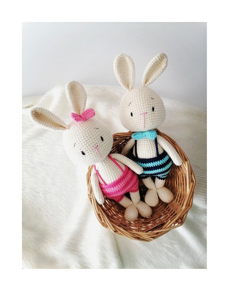 May include: Two crocheted bunny plushies in a wicker basket. The bunny on the left is wearing a pink and white outfit, while the bunny on the right is wearing a blue and white outfit.
