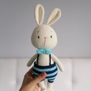 May include: A white crocheted bunny wearing blue and turquoise striped overalls with a turquoise bow tie.