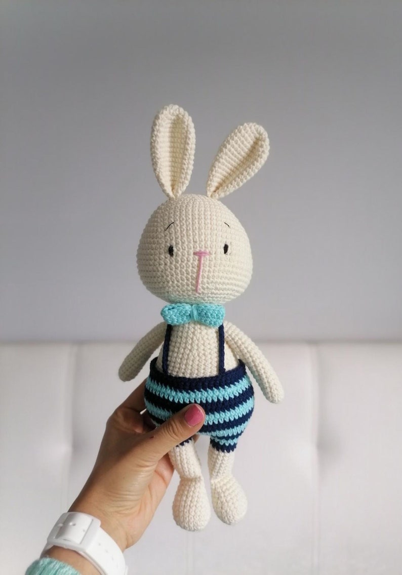 May include: A white crocheted bunny wearing blue and turquoise striped overalls with a turquoise bow tie.