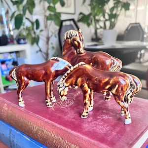 May include: Three brown ceramic horse figurines, two grazing and one reclining, are displayed on a stack of antique books with red and blue covers. The horses have white accents on their manes and hooves.
