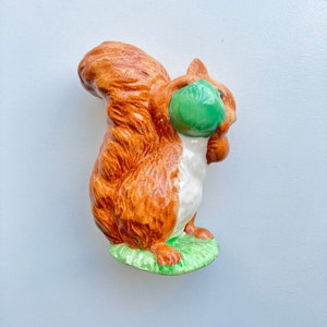May include: A ceramic squirrel figurine with a brown and white body, holding a green acorn in its paws. The squirrel is standing on a green base.