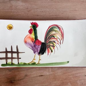 May include: A white ceramic rectangular dish with a hand-painted rooster design. The rooster is purple, red, yellow, and green with a black tail. The dish has a brown fence and a yellow sun.