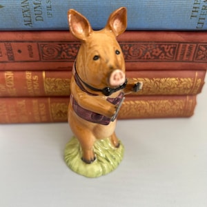 May include: A ceramic pig figurine standing on a green base, wearing a black bow tie and a purple vest. The pig is light brown with pointed ears, a snout, and a small tail. It is depicted in an upright, anthropomorphic pose with its arms extended.