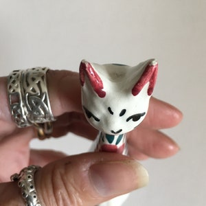 Puede incluir: Figurilla de cer&aacute;mica blanca de un gato con detalles pintados en rojo, azul y verde. El gato tiene una expresi&oacute;n caprichosa y est&aacute; de pie sobre sus patas traseras.