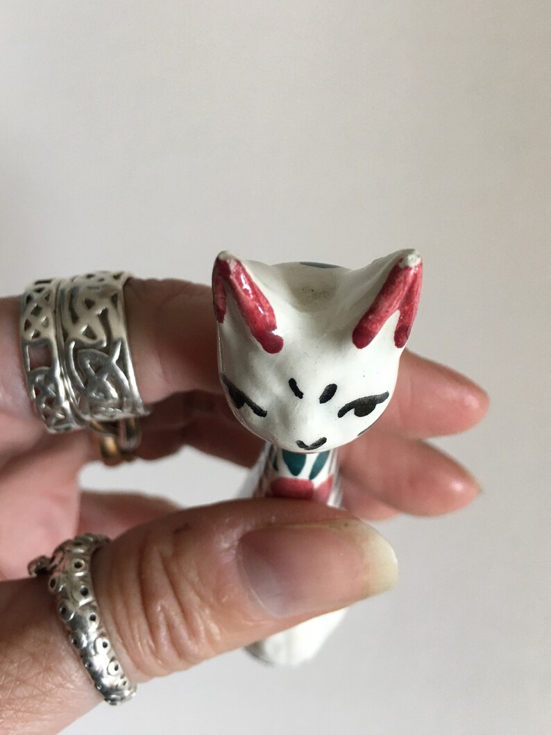 Puede incluir: Figurilla de cer&aacute;mica blanca de un gato con detalles pintados en rojo, azul y verde. El gato tiene una expresi&oacute;n caprichosa y est&aacute; de pie sobre sus patas traseras.