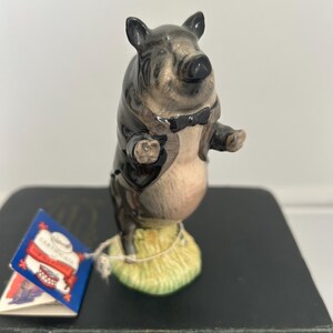 May include: A ceramic pig figurine standing on a textured green base, wearing a black tuxedo jacket and a black bowtie. The pig has a glossy finish with grey and cream coloring. A small blue and white paper tag is attached to the base by a string.