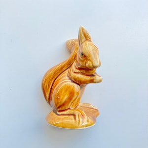 May include: A ceramic squirrel figurine with brown and orange glaze. The squirrel is sitting on a base and has its tail curled around its body.