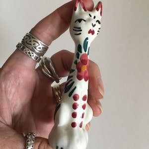 Puede incluir: Una figurita de gato de cer&aacute;mica blanca con detalles pintados en rojo, verde y negro. El gato tiene un cuerpo largo y delgado y un dise&ntilde;o floral en la espalda.