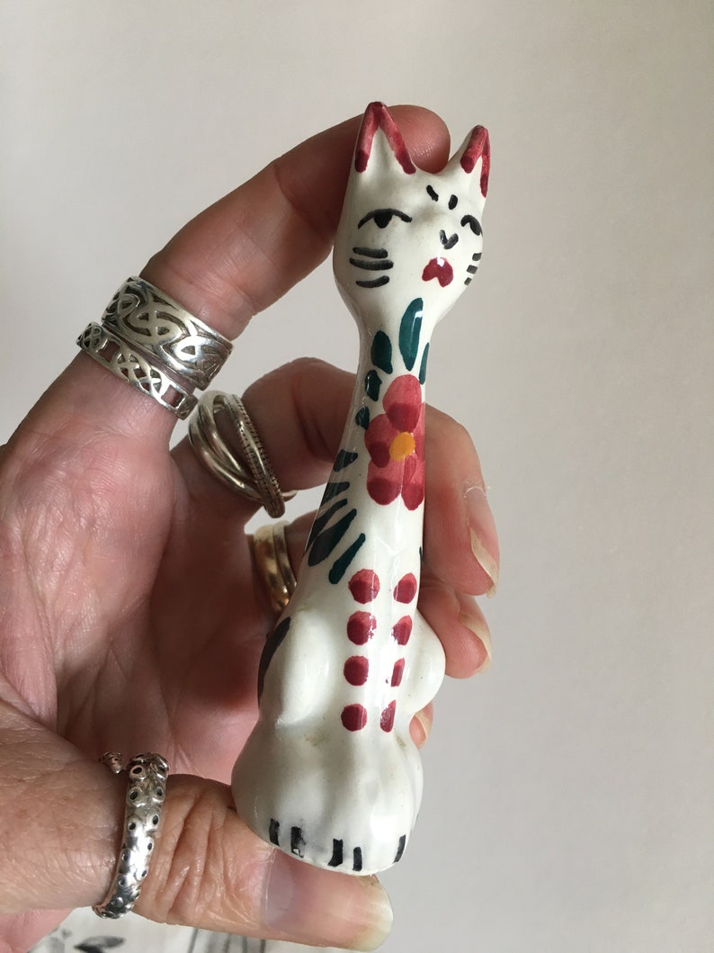Puede incluir: Una figurita de gato de cer&aacute;mica blanca con detalles pintados en rojo, verde y negro. El gato tiene un cuerpo largo y delgado y un dise&ntilde;o floral en la espalda.