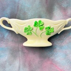 May include: A white ceramic bowl with a green shamrock design. The bowl has two handles and a pedestal base.